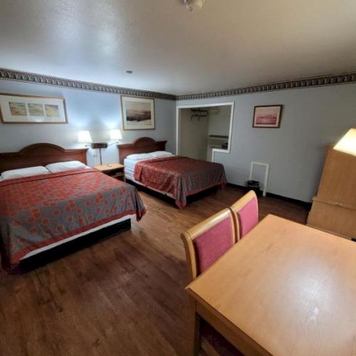 A hotel room with two queen beds, a wooden desk and chair, a dresser with a lamp, and a small seating area, warm lighting, and wood flooring.