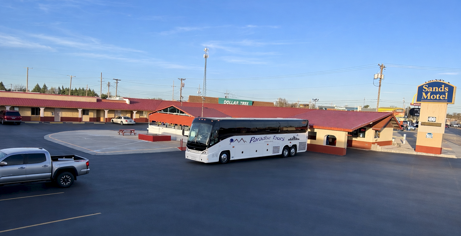 A white tour bus is parked in a desert motel lot near a single-story red-roofed motel with a Sands Motel sign against a blue sky.