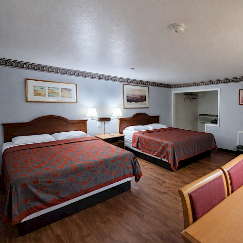 Two full beds in a simple hotel room with a desk, dresser, and a small seating area; warm lighting and wood floors, under a blue-gray wall, with framed pictures.