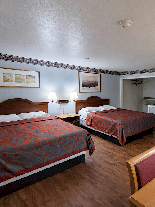 Two full beds in a simple hotel room with a desk, dresser, and a small seating area; warm lighting and wood floors, under a blue-gray wall, with framed pictures.