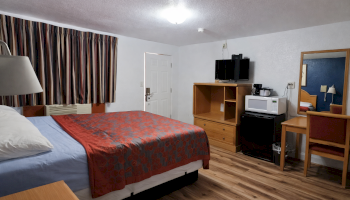 A motel-style room with a queen bed, wooden furniture, TV, microwave, mini-fridge, desk, mirror, and a striped curtain in a cozy, simple setup.