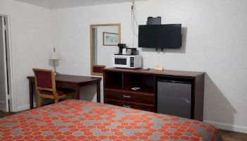 A hotel room setup with a bed in the foreground, a wooden dresser and desk against the far wall, a wall-mounted TV, microwave, mini-fridge, and a desk lamp with a chair, plus a mirror on the wall.