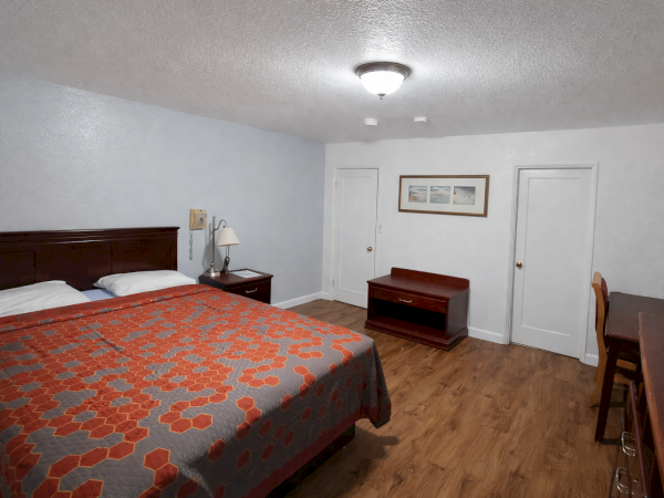 A motel-style hotel room with a large bed, wooden headboard, nightstands, a lamp, two doors, a desk, chair, and a small bench against the wall, laminate flooring.