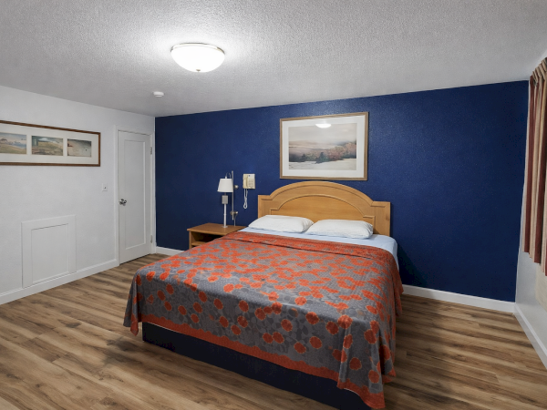 A tidy hotel-style bedroom with a blue accent wall, queen bed, wooden headboard, two nightstands, framed wall art, and a patterned orange-gray bedspread.