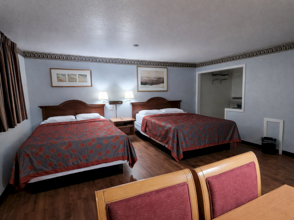 A plain hotel room with two double beds, matching lamps, wall art, a small bench, and a partial view of a table and chairs in the foreground.