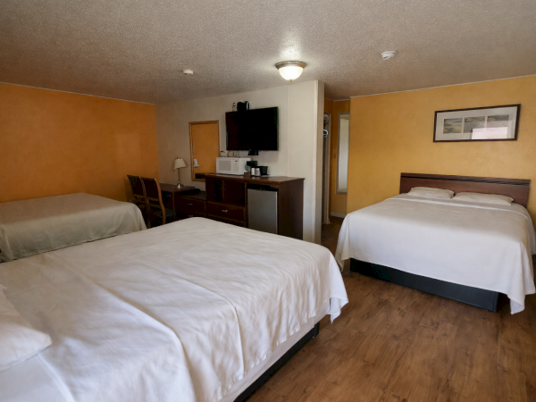 A hotel room with three beds (two queen/king and one extra), a dresser with a mounted TV, a chair, and warm tan walls.