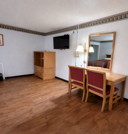 A simple hotel room with a wooden floor, a bed partially shown, a dresser, wall-mounted TV, desk with mirror and two red chairs, and a small framed picture on the wall.