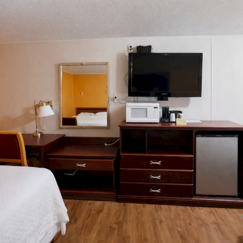 A hotel room with two beds, a desk with a mirror, a mini-fridge, microwave, and a mounted TV on a white accent wall.