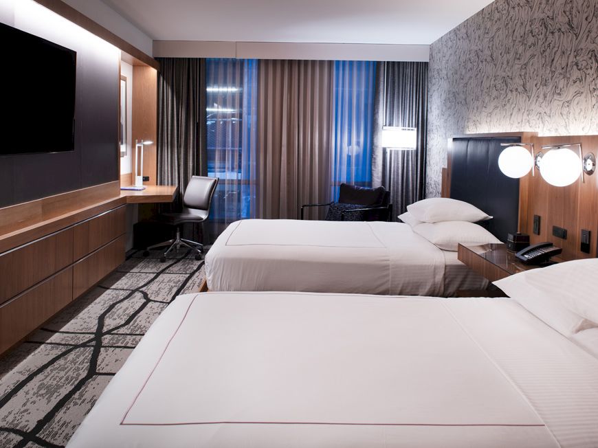 A modern hotel room with two white beds, a wall-mounted TV, desk lamp lights, and dark curtains behind the window.