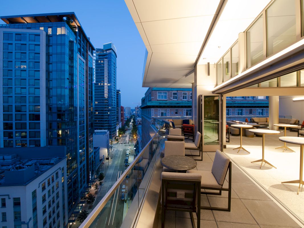 Rooftop balcony with modern lounge seating and tables, looking over a city street between tall buildings.